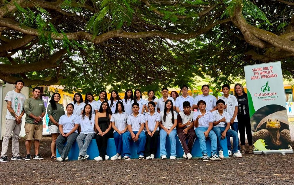 An image of 22 high school students wearing a mix of light and dark colored t-shirts, flanked by Galápagos Conservancy staff members. The Galápagos Conservancy logo and branding stands on a banner on the right hand side of the image.
